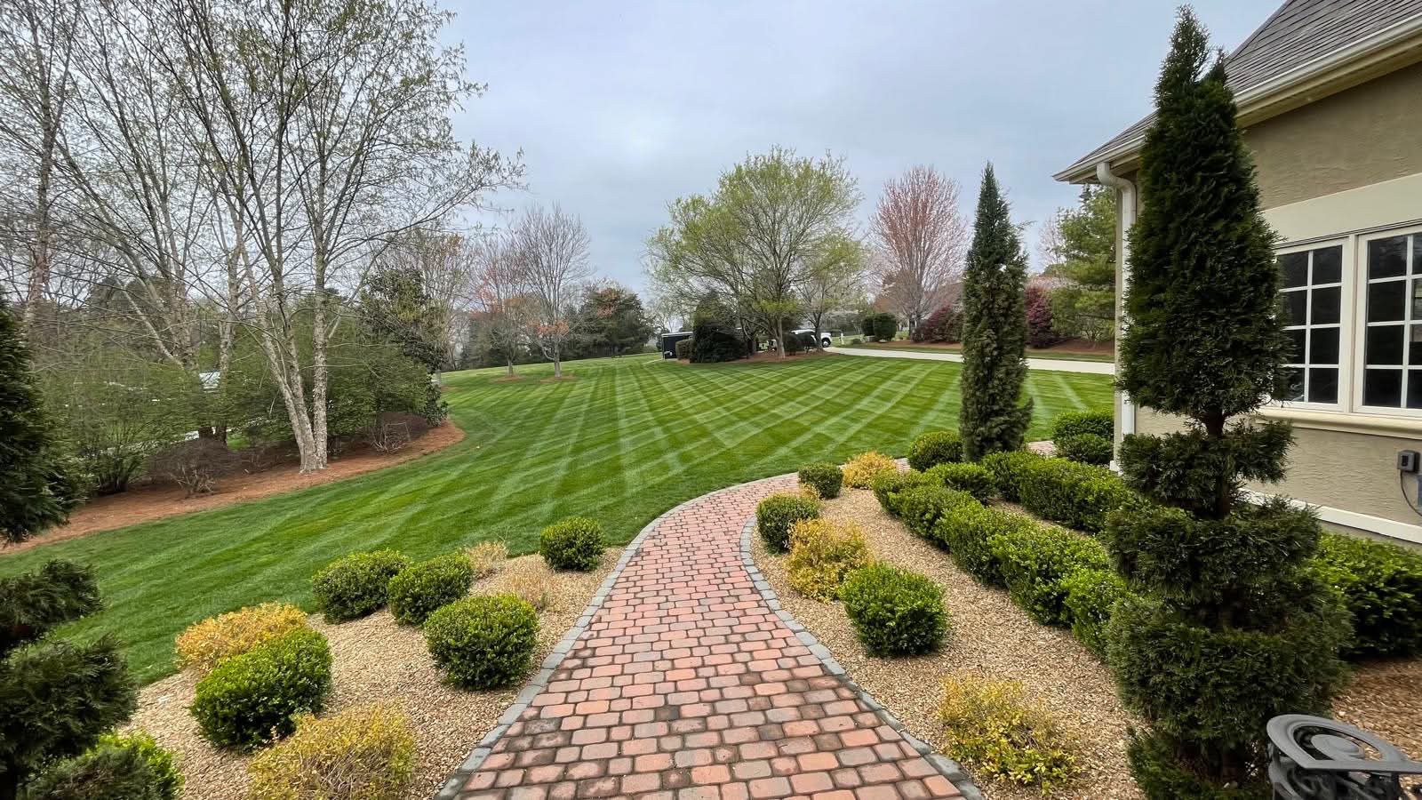A striped green lawn and curved brick walkway through neatly trimmed shrub beds beside a home.